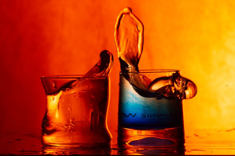 Two glasses with colored liquid splashing dramatically against a vibrant orange background.